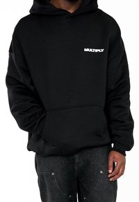 Persona che indossa una felpa nera con cappuccio con il testo "MULTIPLY" sul petto e jeans scuri con grandi tasche frontali, mano appoggiata di lato.