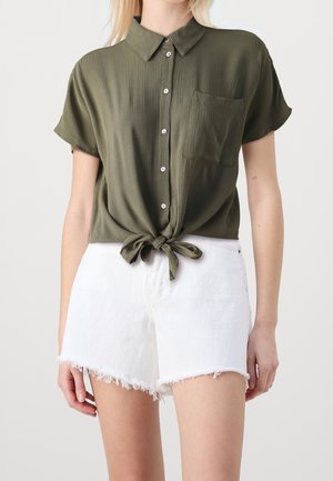 Camicia verde oliva con maniche corte, bottoni e vita annodata, abbinata a pantaloni corti bianchi con orlo sfrangiato.