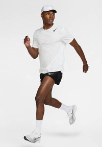 Λευκό κοντομάνικο μπλουζάκι Nike, μαύρο σορτς Nike και γκρι παπούτσια τρεξίματος. Το μοντέλο τρέχει σε φωτεινό περιβάλλον, φοράει λευκό καπέλο και λευκές κάλτσες.