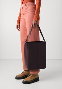 Sac fourre-tout violet foncé avec un design rectangulaire structuré, texture lisse et anses courtes. Associé à un haut orange à manches longues et un jean rose.