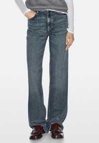 Jean à jambes larges en denim bleu foncé avec une finition légèrement délavée. Comprend cinq poches et une fermeture classique à bouton. Associé à des mocassins marron.