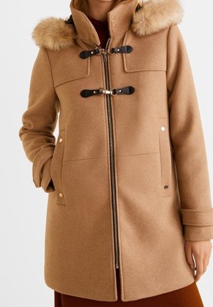 Manteau marron chameau avec capuche bordée de fourrure, deux attaches à bascule noires, fermeture éclair et poches latérales, porté par une personne les mains dans les poches.