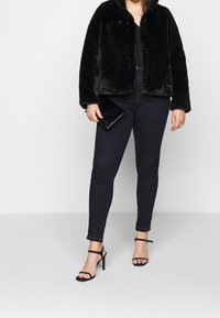 Sort faux fur jakke, cropped design, lange ærmer. Matchet med mørke slim-fit jeans og sorte strap-højhælede sandaler. Holder en sort clutch.