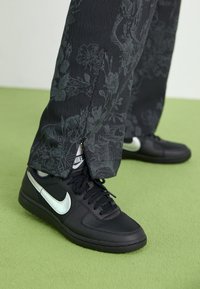 Černé tenisky Nike s bílým logem swoosh nošené s černými kalhotami s květinovým vzorem na zelené podlaze.
