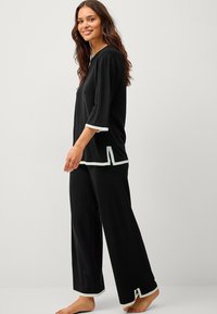 Sort to-parts pyjamasæt med en løs, langærmet top med rund hals og hvid kant, parret med vide bukser med lignende detaljer.