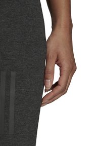 Graue Sportleggings aus dehnbarem Material, mit drei vertikalen schwarzen Streifen an der Seite und einer Nahaufnahme einer Hand, die dagegen ruht.