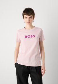 Šviesiai rožinė medvilninė marškinėliai su žodžiu "BOSS" ryškiomis violetinėmis raidėmis. Apvalus kaklas, trumpomis rankovėmis. Modelis derinamas su juodomis kelnėmis.