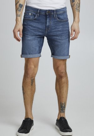 Shorts en denim bleu foncé avec une coupe classique, présentant des revers et cinq poches. Portés avec des baskets noires.
