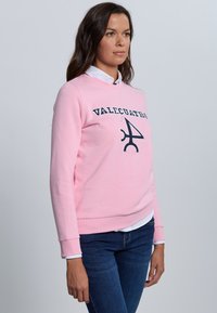 Sudadera rosa con cuello redondo con texto bordado "VALCUATRO" y un logo, con puños y dobladillo acanalados, llevada sobre una camisa blanca con cuello.