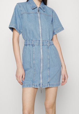 Mujer con un vestido corto de manga corta, de mezclilla azul claro, con cremallera frontal y cintura elástica, posando frente a un fondo blanco.