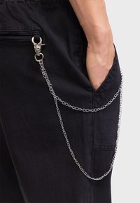 Pantalones de tela negra con un bolsillo lateral, un accesorio de cadena de metal plateado con broche, y un acabado texturizado.