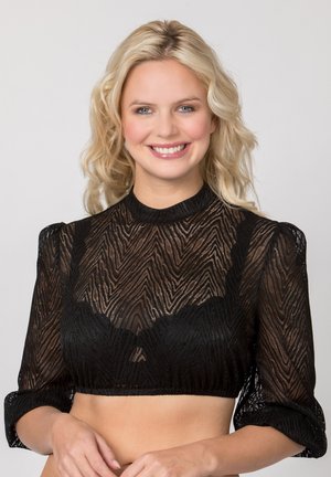 Blonde Frau lächelt, trägt ein durchsichtiges schwarzes gemustertes Crop-Top mit Dreiviertelärmeln vor einem schlichten hellen Hintergrund.