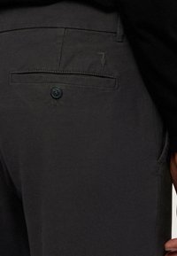 Tmavě šedé kalhoty mají zadní kapsu s knoflíkovým zavíráním, decentní logo a textilní strukturu.