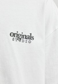 T-shirt bianco in cotone con testo nero "originals STUDIO", dalla vestibilità comoda e maniche corte. Il testo è leggermente fuori centro.