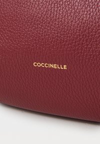Sac à main en cuir bordeaux avec une surface texturée et un logo "Coccinelle" doré embossé près du bas.