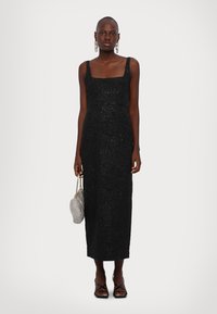 Schwarzes Midikleid mit quadratischem Ausschnitt, strukturiertem Stoff, taillierter Silhouette und breiten Trägern. Kombiniert mit einer silbernen Handtasche und schlichten Schuhen.