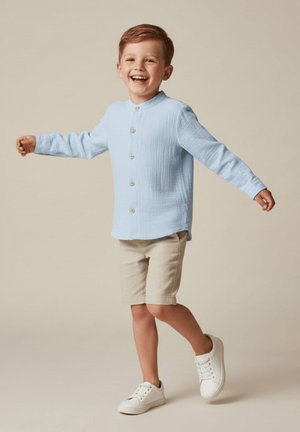 Uśmiechnięty młody chłopiec w jasnoniebieskiej koszuli z guzikami, beżowych szortach i białych trampkach, stoi z wyciągniętymi ramionami na neutralnym tle.