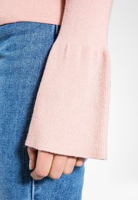 Manche de pull côtelé rose clair, présentant un design évasé, associé à un jean en denim bleu, mettant en avant un contraste de textures décontractées.