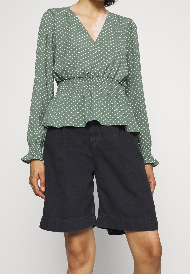 Blusa peplum verde con pois bianchi, maniche lunghe e scollatura a V, abbinata a pantaloni bermuda in denim scuro. Tessuto texturizzato con vita elastica.