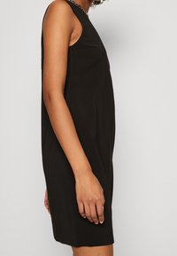 Sødsløs sort kjole med en lige silhuet. Fremstillet af blødt stof har den et enkelt design og en kædeaccent ved halsudskæringen.