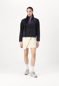 Svart fleece-pullover med abstrakt mönster, halvdragkedja fram och lila accenter, parat med en beige kjol och ljusa sneakers.