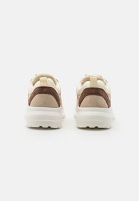 Paire de baskets beige et marron avec des semelles blanches épaisses vues de dos sur un fond clair uni.