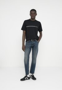 Μαύρο κοντομάνικο μπλουζάκι με λευκή οριζόντια ρίγα, συνδυασμένο με ξεβαμμένο μπλε τζιν ίσιας γραμμής και μαύρα αθλητικά παπούτσια με λευκές λεπτομέρειες.