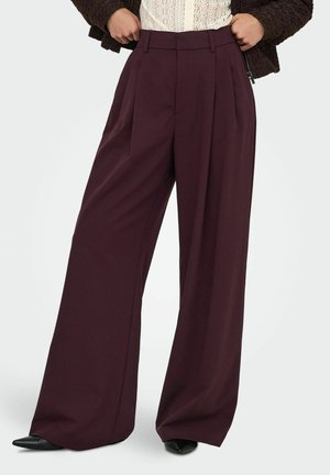 Pantalon ample taille haute à jambes larges de couleur violet foncé, porté avec des chaussures pointues noires et un top en dentelle partiellement couvert par une veste sombre.