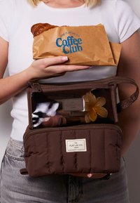 Borsa cosmetica marrone trapuntata con zip, parte superiore trasparente e logo. Una mano tiene una borsa da caffè con un dolce. Contiene articoli di bellezza.