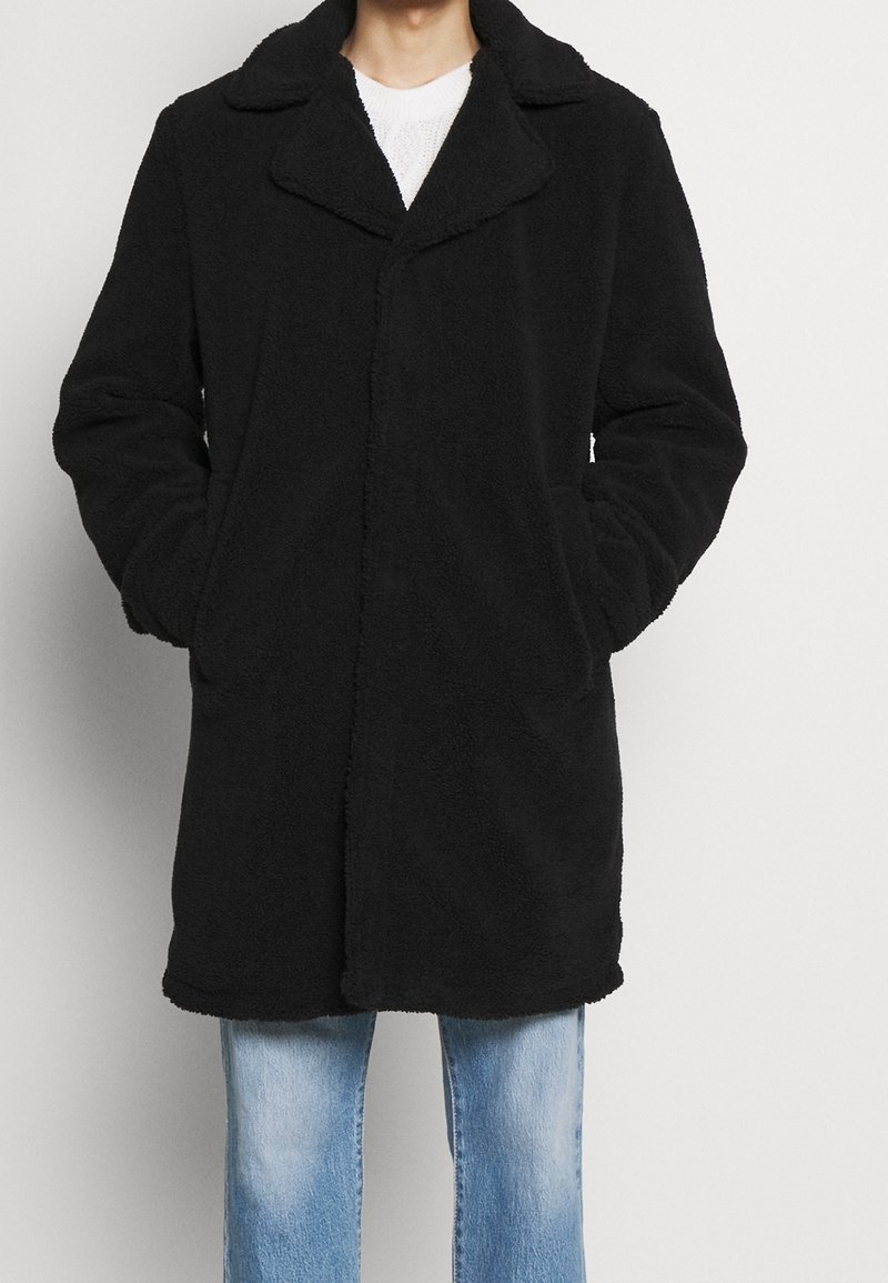Manteau en polaire noire avec col, coupe droite, manches longues et poches latérales, associé à un jean bleu clair.