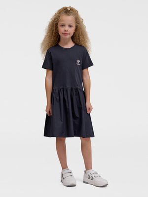 Młoda dziewczyna z kręconymi blond włosami, ubrana w czarną sukienkę z krótkim rękawem i białe trampki, stoi na jednolitym tle.