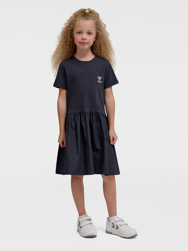 Junges Mädchen mit lockigem blondem Haar, das ein schwarzes kurzärmeliges Kleid und weiße Sneakers trägt, steht vor einem schlichten Hintergrund.