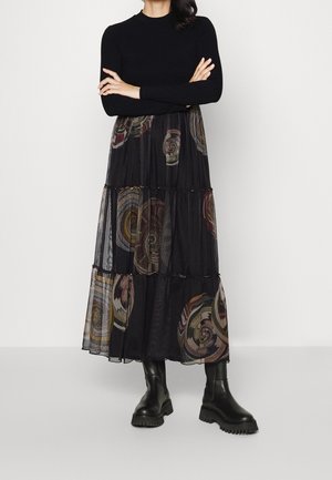 Vrouw draagt een zwart top met lange mouwen, een zwarte doorschijnende gelaagde rok met cirkelpatronen en zwarte chunky laarzen, staand met gekruiste armen.