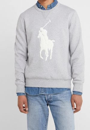 Hombre con sudadera gris con logo blanco de jugador de polo, camisa de mezclilla azul debajo y vaqueros azul claro.
