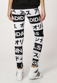 Osoba v černo-bílých legínách Adidas s japonskými znaky a bílými vysokými teniskami Adidas, stojící s otočeným zády.