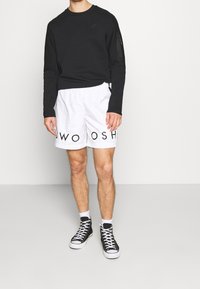 Musta pitkähihainen collegepaita, jossa vetoketjullinen tasku, yhdistettynä valkoisiin shortseihin, joissa musta "SWOOSH"-kuvio. Päällä mustat korkeavartiset tennarit.