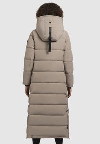 Abrigo puffer beige con un diseño largo y acolchado, capucha ajustada y cremalleras laterales para ventilación. Tejido texturizado con un acabado mate.