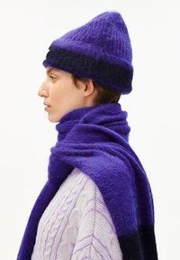 Jeune personne de profil portant un bonnet tricoté violet et noir et une écharpe en peluche violette assortie par-dessus un pull en maille torsadée violet clair.