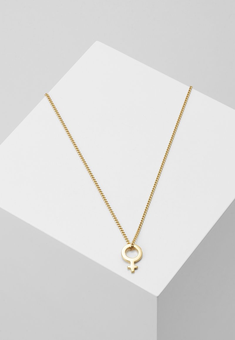 Collier chaîne en or avec un pendentif symbole du genre féminin exposé sur une surface géométrique blanche sur fond gris.