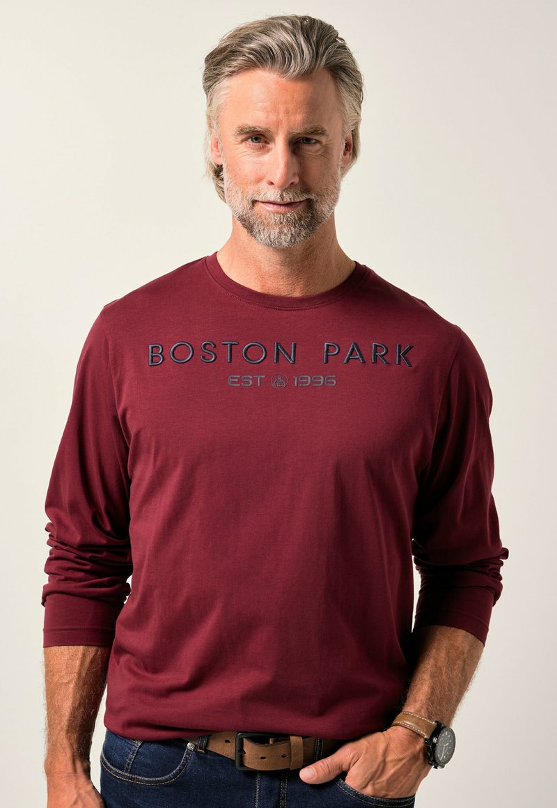 Een man van middelbare leeftijd met grijs haar en een baard, gekleed in een bordeauxkleurig shirt met lange mouwen met de tekst "BOSTON PARK EST 1996", blauwe jeans en een horloge.