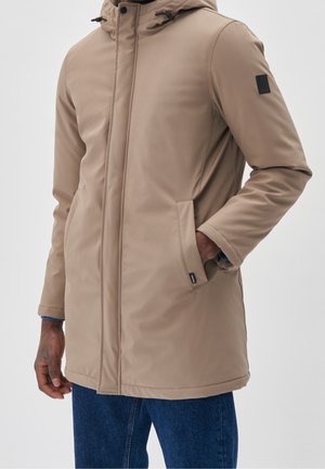 Personne portant un manteau d'hiver beige à capuche avec une main dans la poche, associé à un jean bleu sur un fond uni clair.
