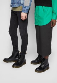 Bottes de combat en cuir noir avec des coutures jaunes, associées à un pantalon slim noir et une chemise verte à boutons, présentées à côté d'une longue jupe foncée.