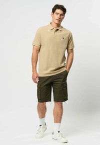 Mladý muž stojící v béžovém polo tričku, tmavě zelených cargo šortkách, bílých ponožkách a bílých teniskách na jednotném světlém pozadí.