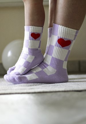 Füße mit lila-weiß karierten Socken mit roten Herzen, drinnen auf einem Teppich neben einem weißen runden Gegenstand.
