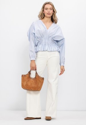 Vrouw draagt een lichtblauw gerimpeld blouse, witte wijde broek, bruine schoenen, en houdt een bruine gevlochten leren handtas vast tegen een witte achtergrond.