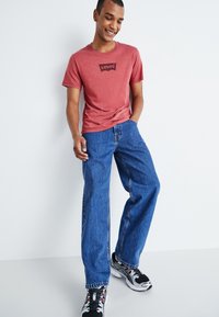 T-shirt vermelha de manga curta com o logótipo preto da Levi's, combinada com jeans azul denim de corte solto e sapatilhas atléticas pretas. Fundo simples.