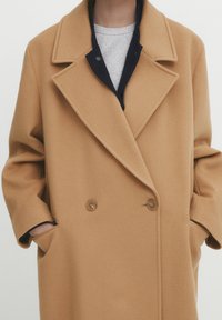 Manteau en laine mélangée couleur chameau avec col cranté, devant croisé à double boutonnage et poches latérales. Doublure matelassée bleu marine visible au niveau du col.