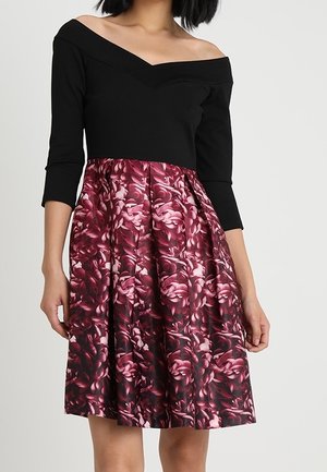 Vrouw draagt een zwarte off-shoulder jurk met driekwart mouwen en een geplooide rok met een roze en donker bloemmotief.