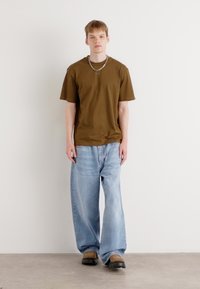 Braunes Baumwoll-T-Shirt, lockere blaue Jeans und schwarze Schuhe. Einfaches Design mit lässigem Schnitt und metallischem Kettenaccessoire.