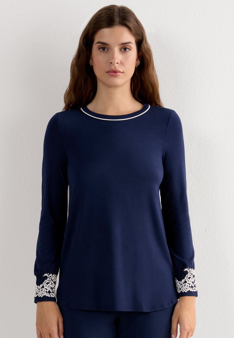 Mujer con una camiseta azul marino de manga larga con detalle de encaje blanco en los puños y un fino ribete blanco alrededor del escote redondo, de pie junto a una pared blanca.